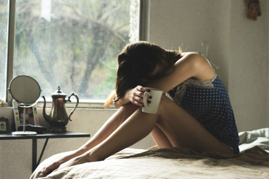 woman sitting on bed with head on knees holding coffee