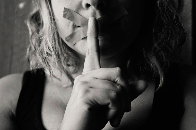 woman with tape over moth and finger to mouth in shush motion